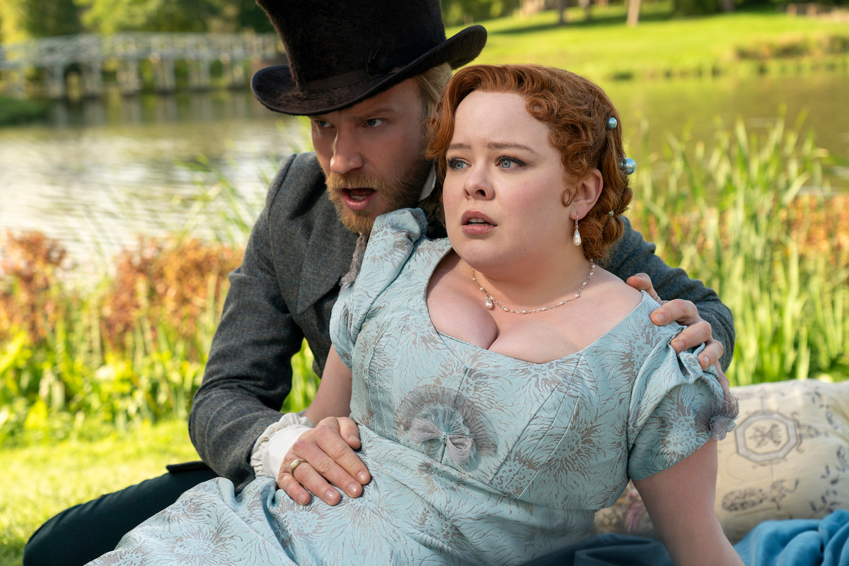 Two people in Regency-era clothing sit close together on a picnic blanket by a scenic lake, both appearing surprised or alarmed, with lush green grass and a bridge visible in the background.