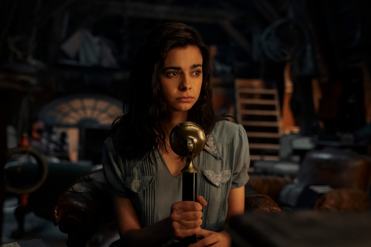 Marie-Laure (played by Aria Mia Loberti) is in a dimly lit room holding a radio broadcast microphone in front of her. She has dark hair and eyes and fair skin and is wearing a light blue, short-sleeved dress.