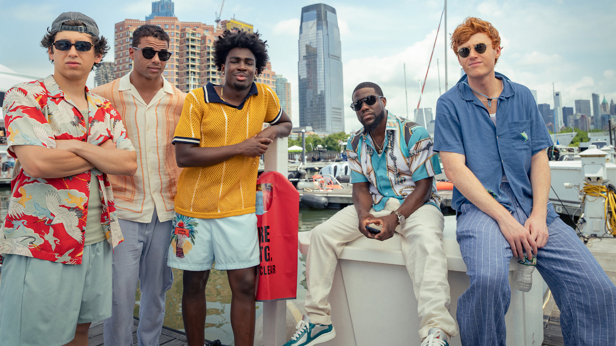 Five young men in casual summer outfits stand by a marina with boats, modern city buildings, and a waterfront skyline in the background, suggesting a relaxed and urban environment.