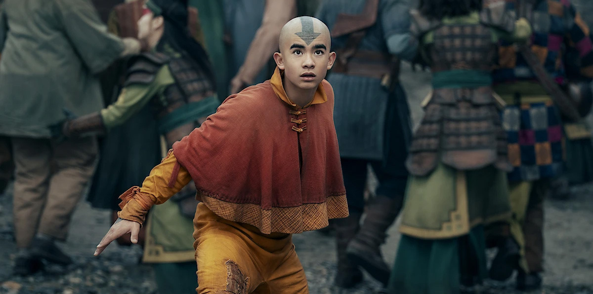 A young bald person with an arrow tattoo, dressed in orange and yellow robes, stands alert in a crowded, action-filled outdoor setting with people in armor and traditional clothing.