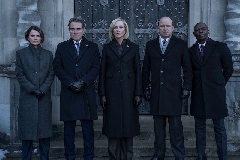 Five people wearing dark formal coats and gloves stand side by side in front of an ornate, heavy wooden door, looking serious and composed.