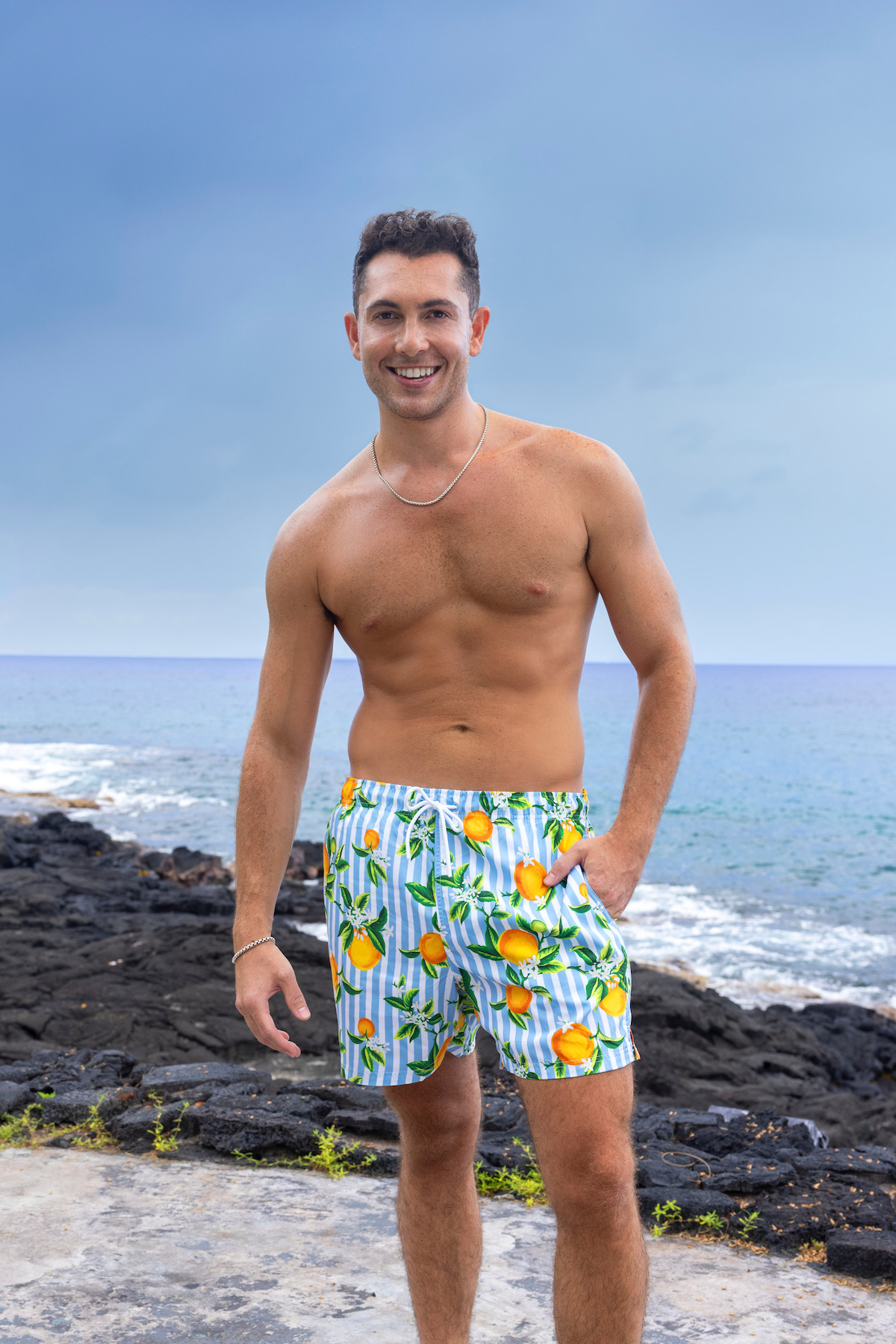 Shirtless man in colorful swim trunks stands on rocky beach with ocean waves and cloudy blue sky in the background.
