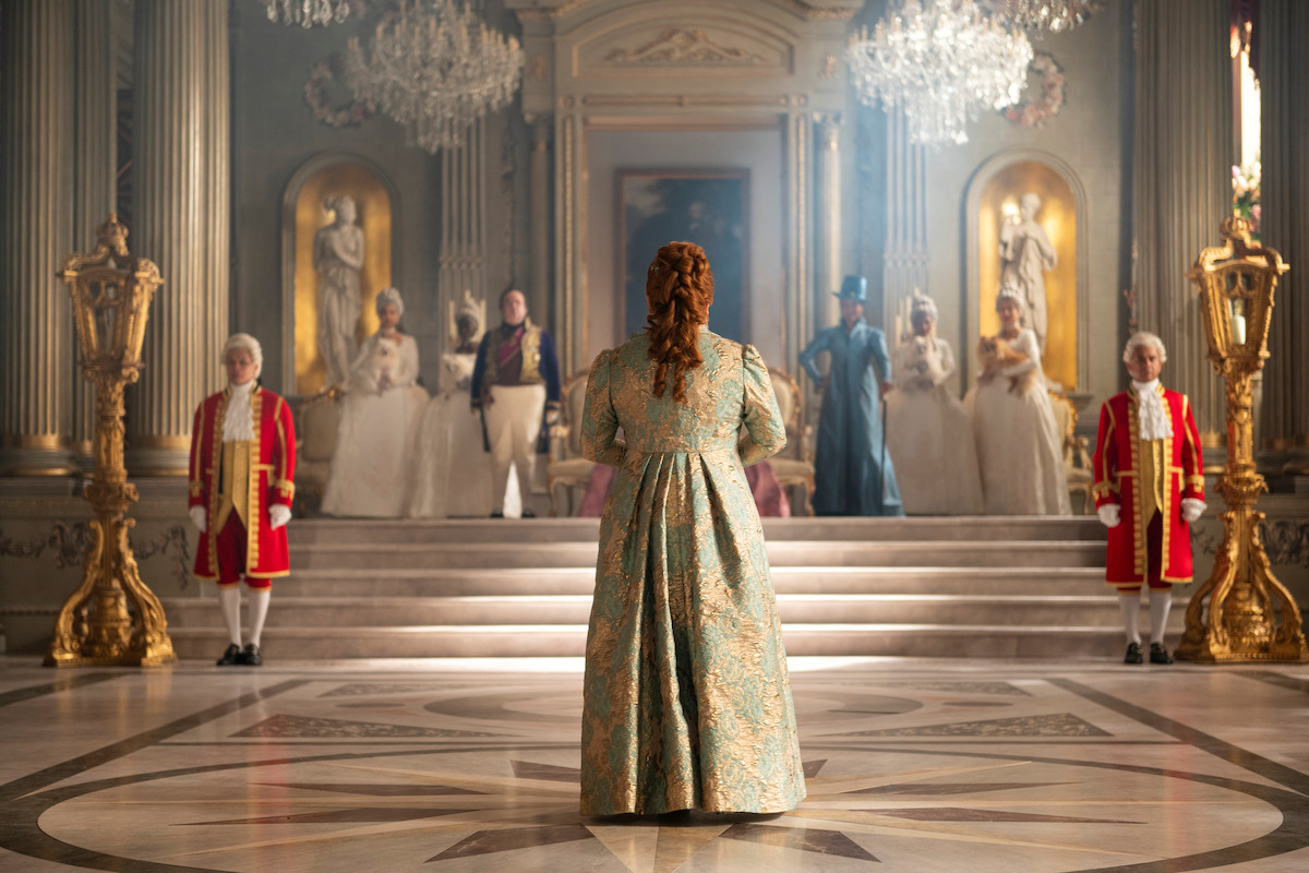 A woman in an ornate gown stands in a grand, elegant hall facing a group in historical costumes on a staircase, surrounded by chandeliers and gold decor, evoking a royal or aristocratic setting.