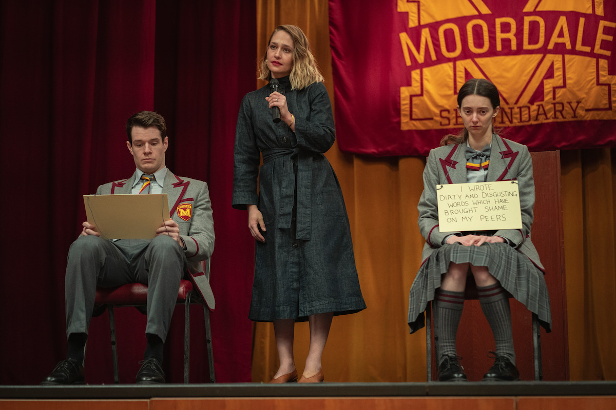 Connor Swindells as Adam Groff and Tanya Reynolds as Lily Iglehart sit on the stage of an auditorium while Jemima Kirke as Hope Haddon stands between them holding a microphone in Season 3 of ‘Sex Education.’