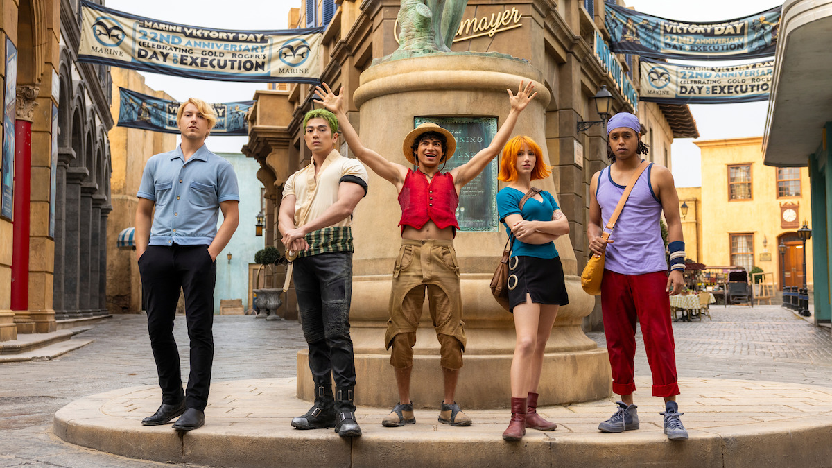 Five people in colorful costumes pose in front of a statue in a lively, detailed town square with banners overhead, suggesting a festive or adventurous atmosphere.