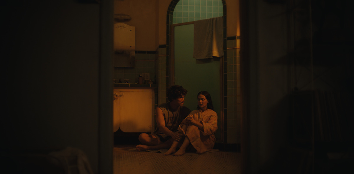 Two people sit on a bathroom floor together at night, illuminated by warm, dim light. The setting is intimate and quiet, with tiled walls, a towel hanging, and a bathtub visible, creating a calm, contemplative mood.