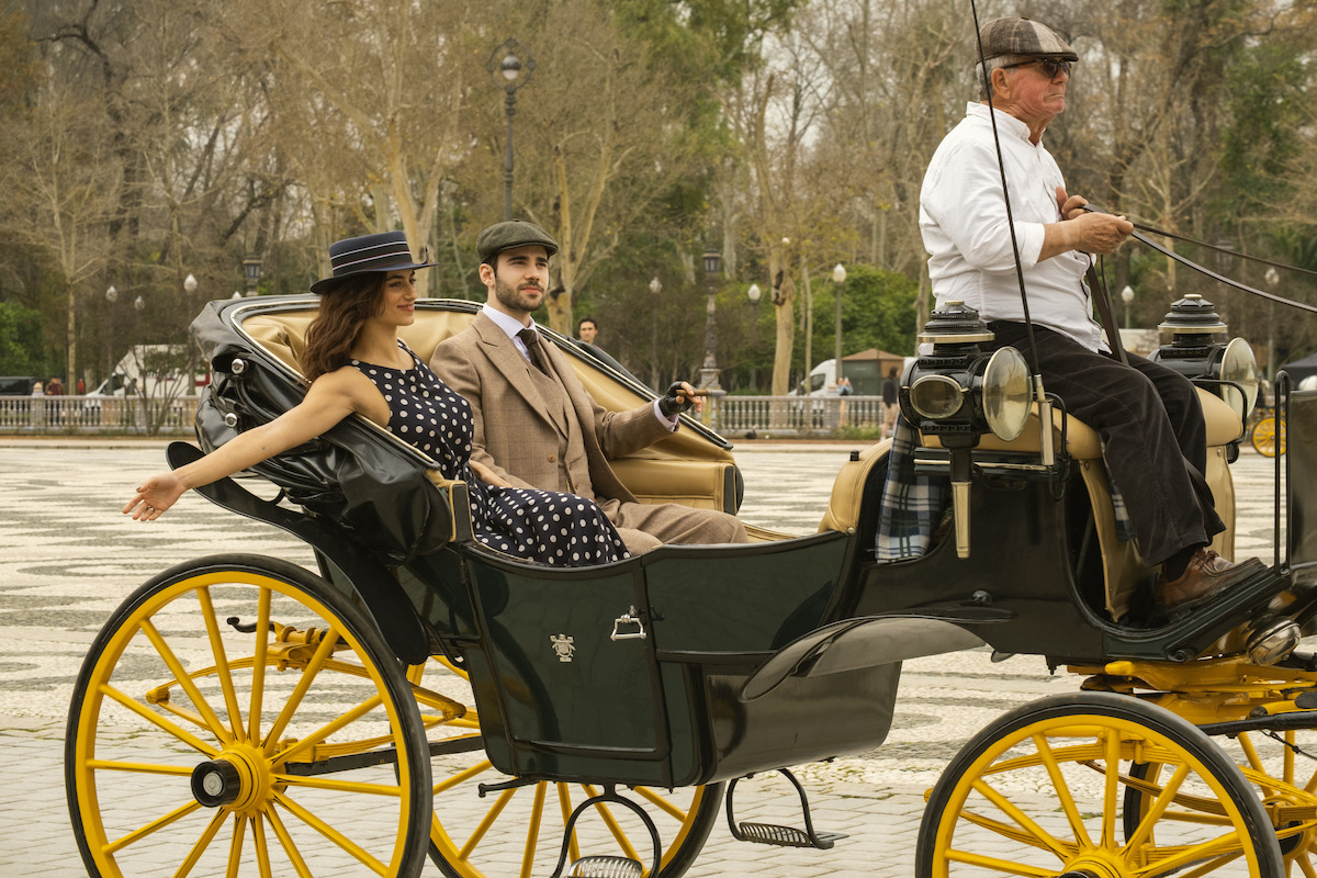 A couple in vintage clothing sits in a black and yellow horse-drawn carriage driven by a man, traveling through a tree-lined park with paved walkways, on a cloudy day.