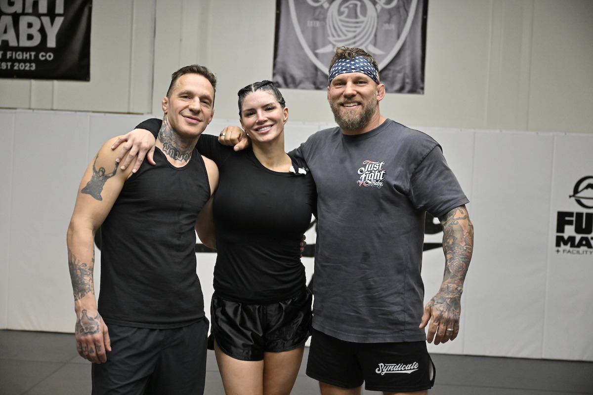Three people in athletic wear standing together, smiling, in a martial arts or gym training facility with banners and mats visible in the background.
