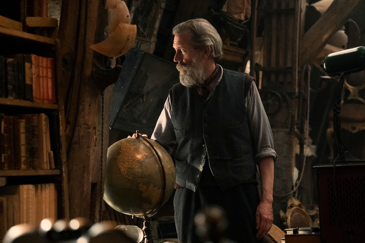 Uncle Etienne (played by Hugh Laurie) stands gazing out an open attic window. He is an older man with gray hair, a gray beard and blue eyes. He wears a light gray long-sleeve shirt with the sleeves rolled up to his elbows with a red scarf, dark gray vest, and dark colored pants. There is a bookcase in the foreground of the cluttered, dimly lit room.