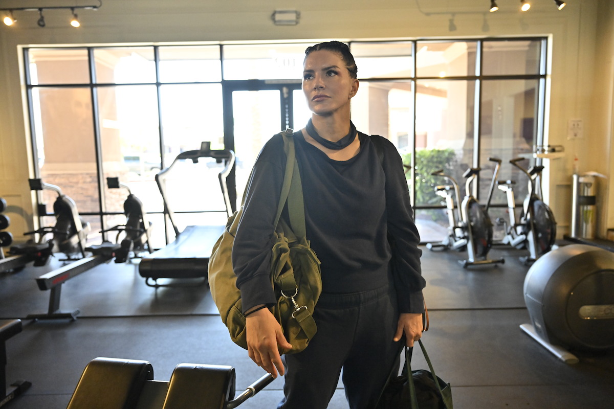 Woman standing in a gym with gym bags, wearing athletic clothes. She is looking forward with exercise equipment, stationary bikes, and weights visible in the background, natural light coming through large glass windows.