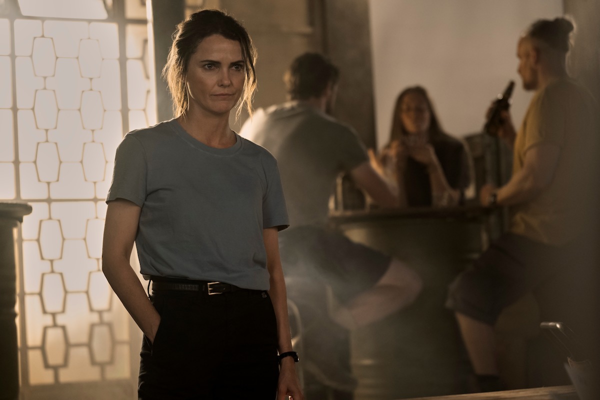 Woman in casual clothes stands in a dimly lit bar or lounge with hands in pockets, looking serious; three people are talking and drinking in the background near a window with patterned glass.