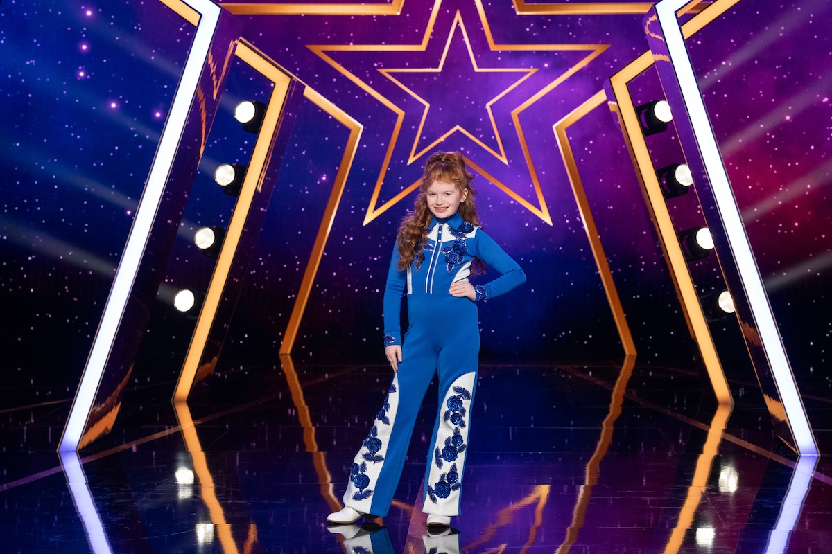 Blair Kudelka in a blue jumpsuit stands confidently on a shiny stage with a large star backdrop, surrounded by bright lights and a colorful, cosmic-themed background.