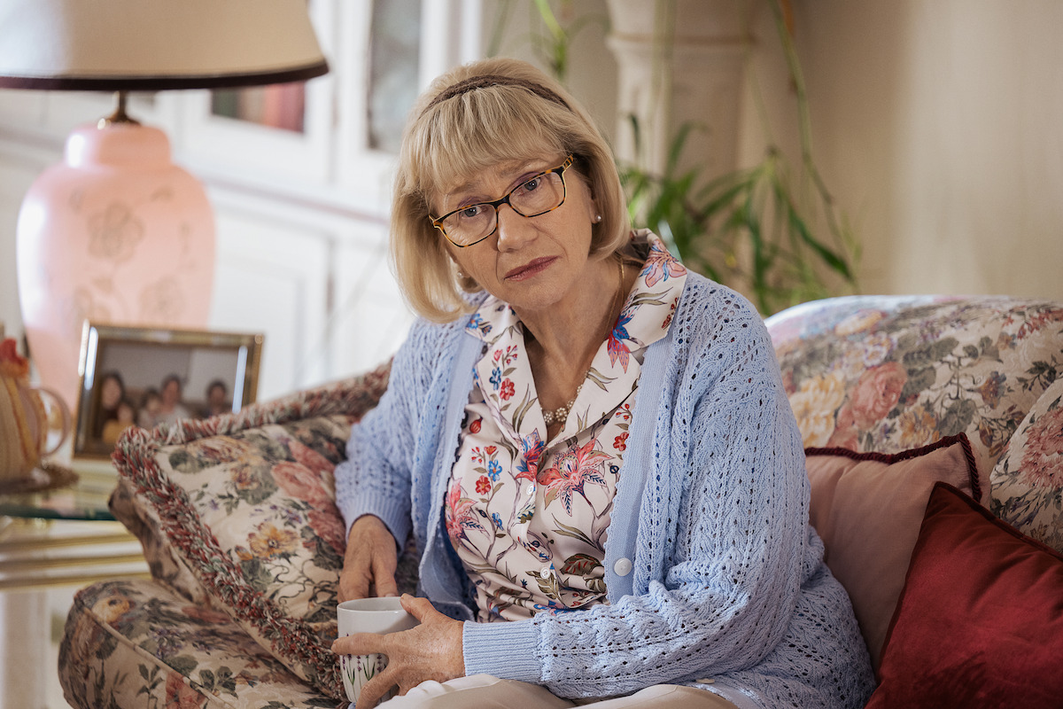 Kathy Baker as Mary Ann, with glasses and floral blouse sits on a floral sofa, holding a mug in a cozy living room with soft light, a pink lamp, framed photo, and plants in the background.