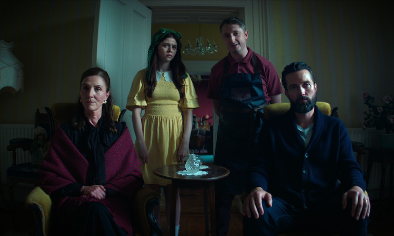 Four people in a dimly lit vintage room, two seated and two standing behind them. The mood is tense and somber, with muted colors and formal, old-fashioned clothing, suggesting a serious or dramatic moment.
