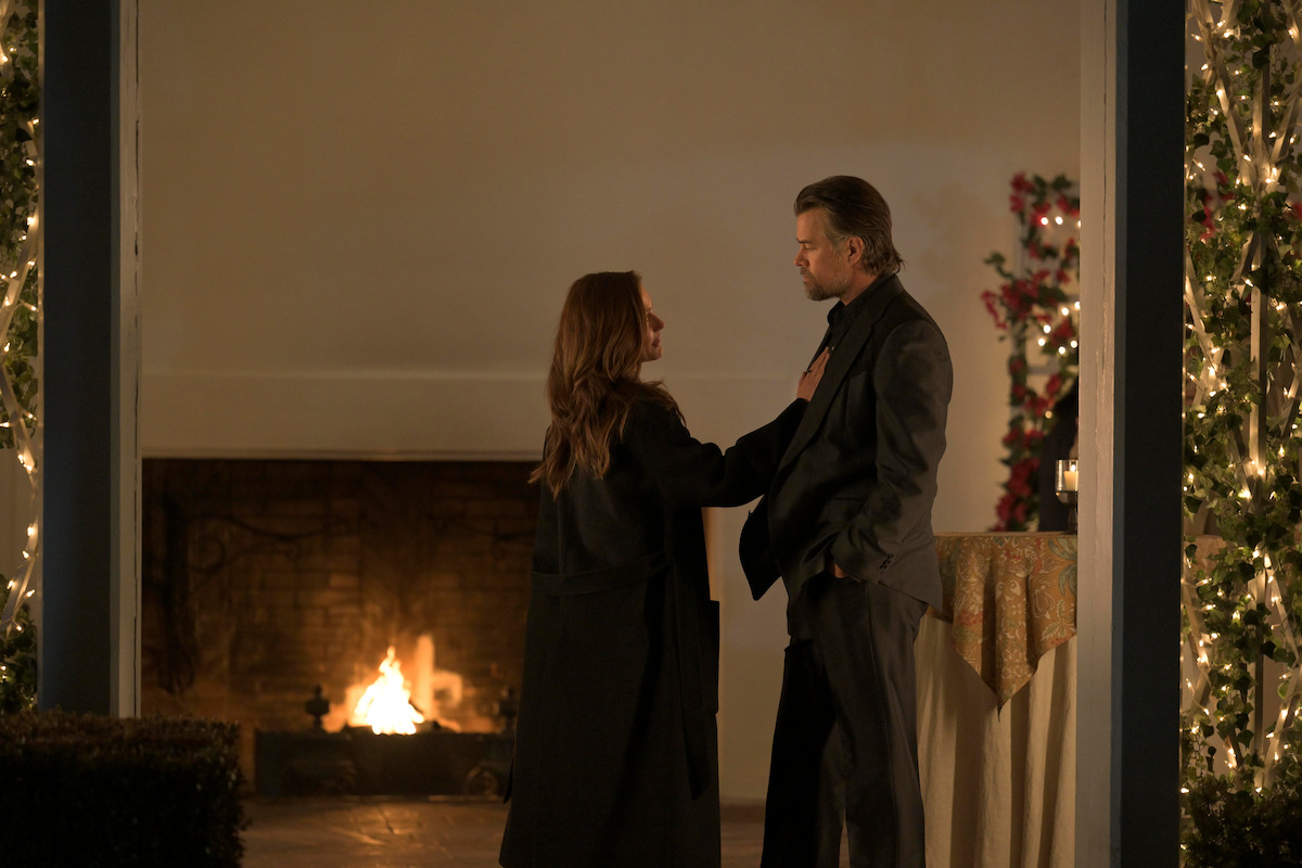 A woman and a man stand facing each other on a decorated patio at night, with string lights, a fireplace burning in the background, and a table covered with a cloth nearby, creating a warm, intimate atmosphere.