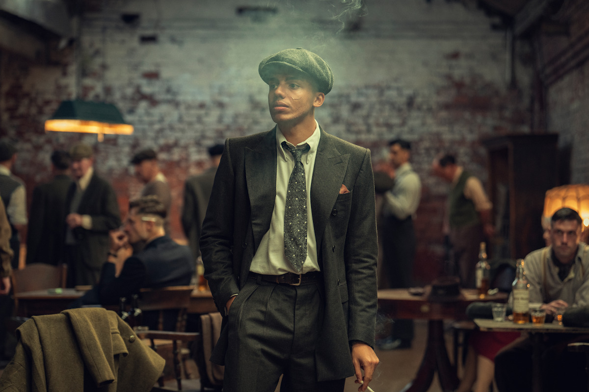 A man in vintage 1920s attire stands in a dimly lit room filled with other men in period clothing, suggesting a social or club setting with a rustic, industrial background.