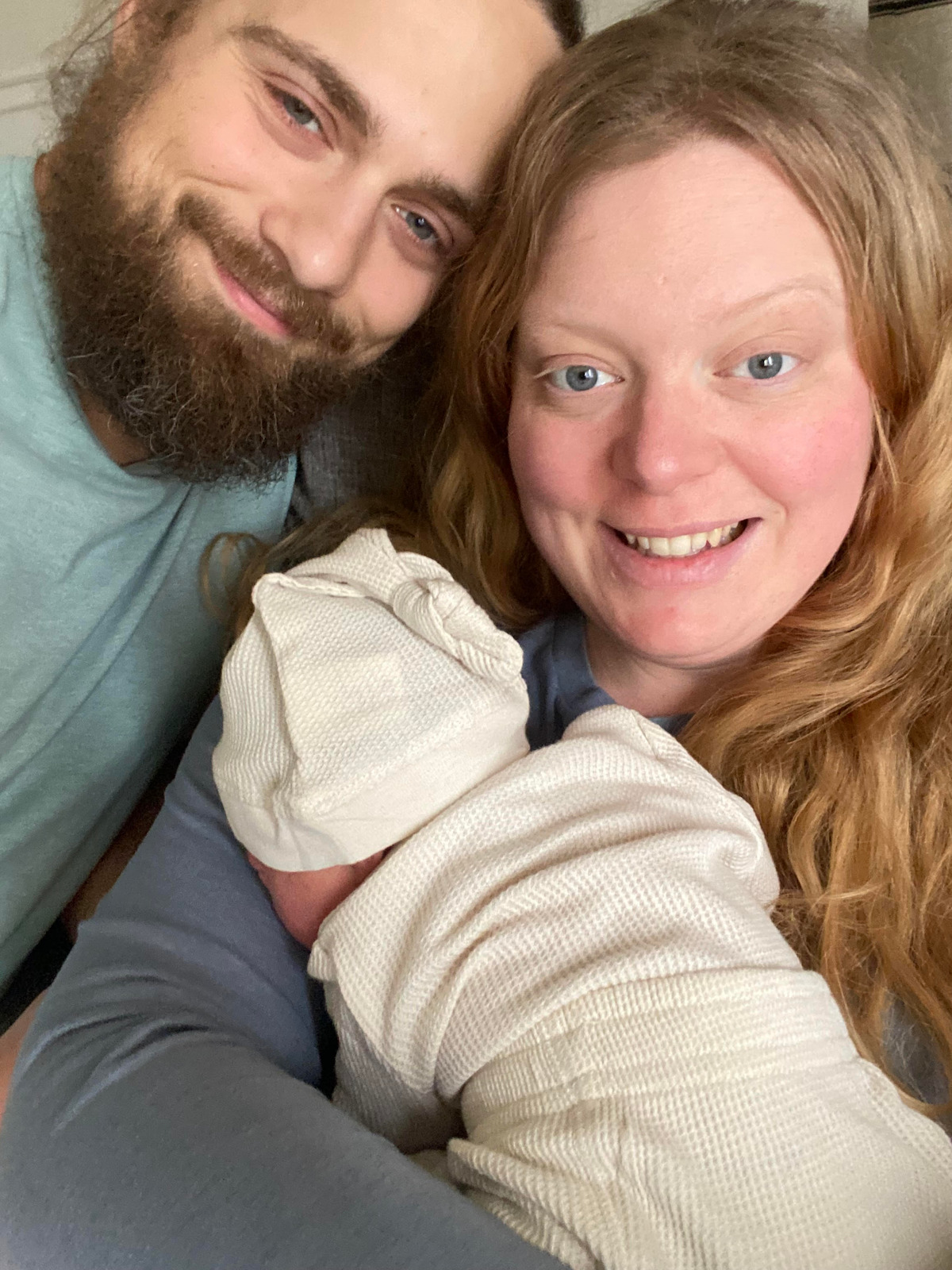 Smiling couple indoors holding a newborn baby wrapped in a cream-colored blanket, capturing a joyful family moment in a close-up setting.