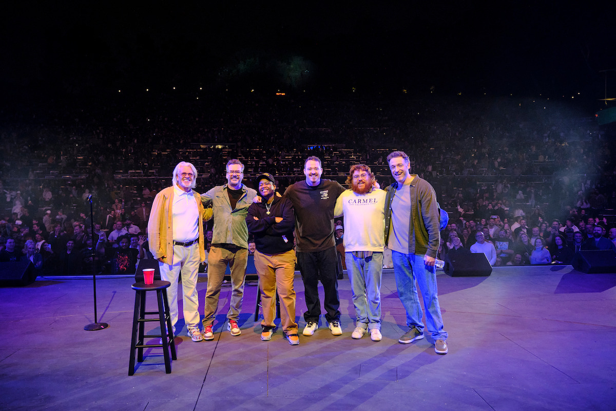 Six men stand together on a stage with a microphone and stool, smiling at the camera as a large audience watches from the darkened outdoor amphitheater behind them at night.