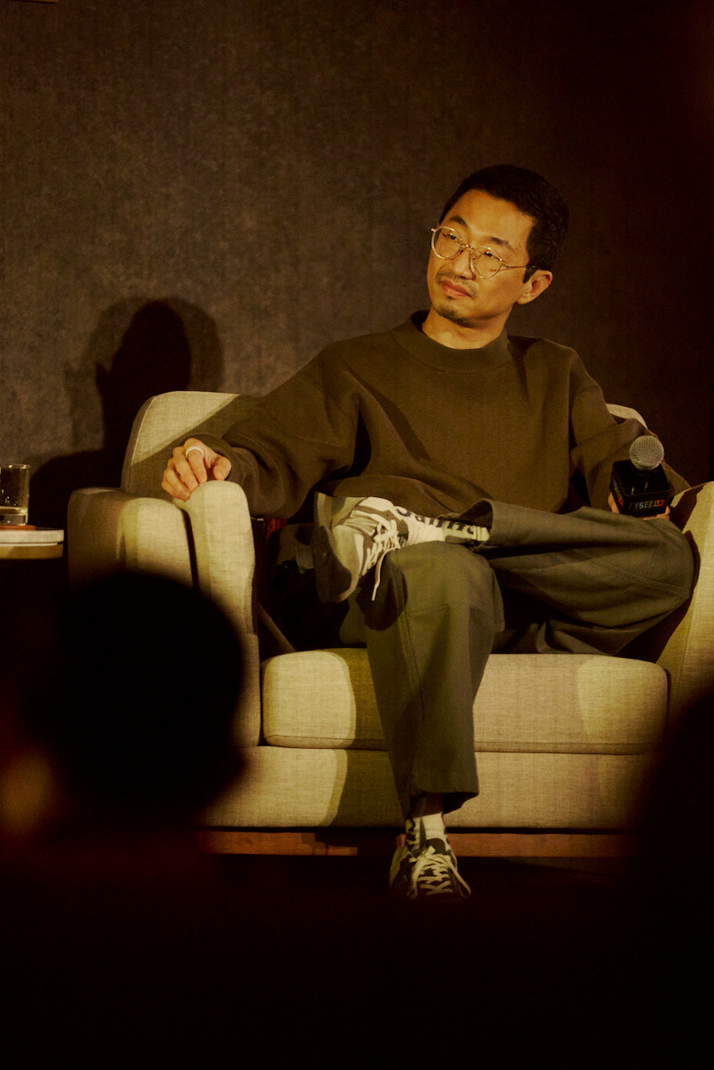 A man wearing glasses and casual clothing sits on a beige armchair on stage, holding a microphone. The background is dark and soft lighting highlights him, suggesting a panel discussion or interview setting.