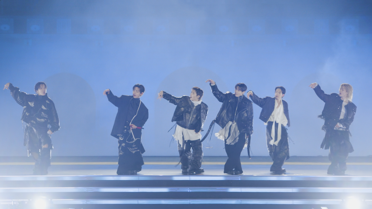 BTS in coordinated dark outfits dance on a brightly lit stage with fog and blue lighting, extending their arms in sync, creating a dynamic and atmospheric concert environment.