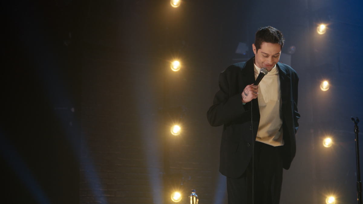 Comedian on stage in a dark suit holding a microphone, performing under warm stage lights set against a brick wall, creating a focused and intimate stand-up comedy atmosphere.