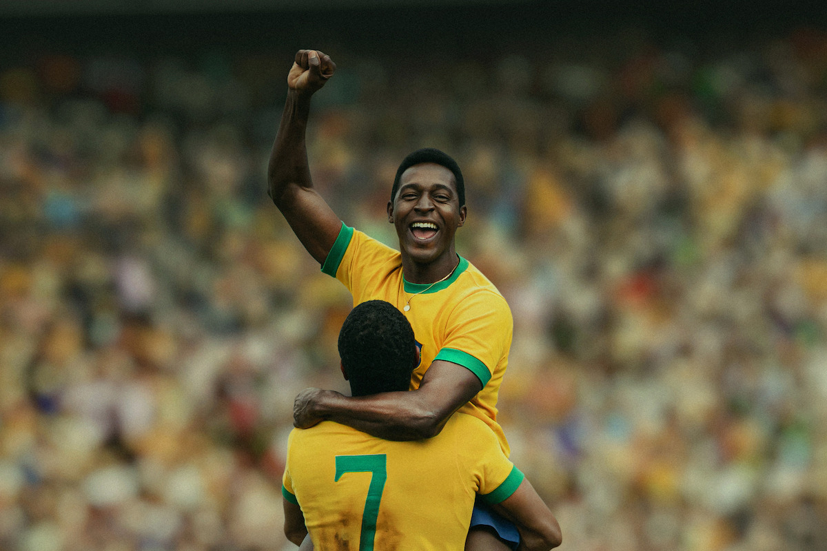 Two soccer players in yellow jerseys celebrate on the field, one raising his fist joyfully while being lifted by a teammate. The stadium background is filled with cheering fans, creating a lively sporting atmosphere.