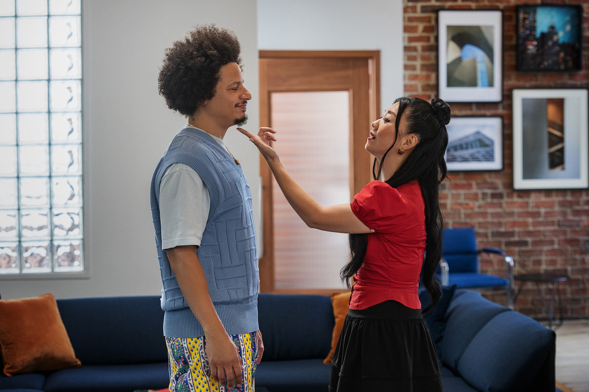 A man and woman stand facing each other in a modern living room with brick walls, framed photos, blue sofa, and orange pillows. The woman is touching the man's chin, both appearing to interact emotionally.