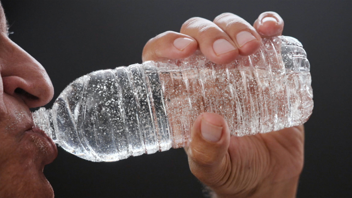 Person drinking water from a clear plastic bottle, held close to their mouth with hand visible, in a close-up shot against a dark background.