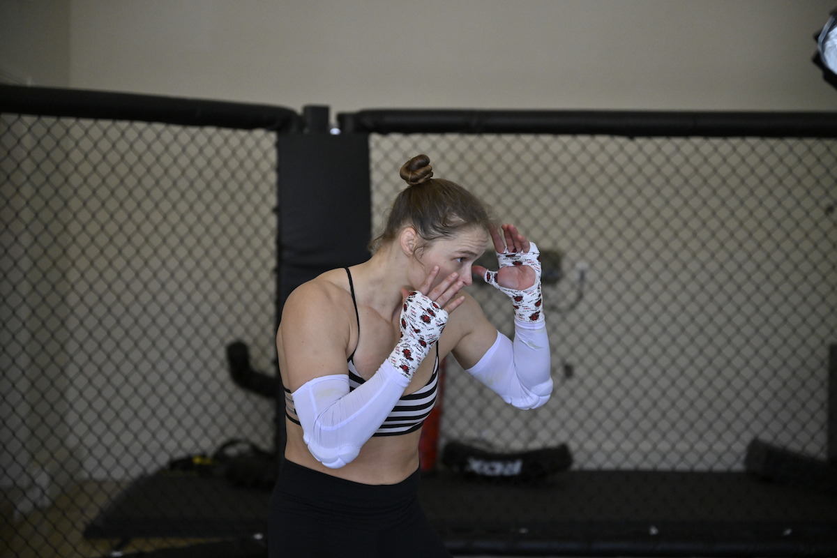Ronda Rousey and Kehlani laughing together in an MMA training gym, standing on mats near a fenced cage, wearing athletic workout clothing with one sporting visible tattoos.