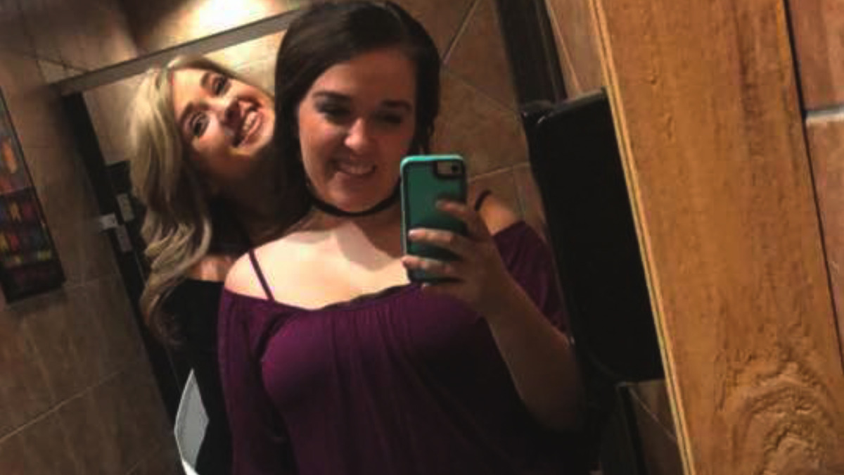 Two women smiling and posing for a selfie in a public restroom with tiled walls and wooden accents, one holding a smartphone in front of a mirror.