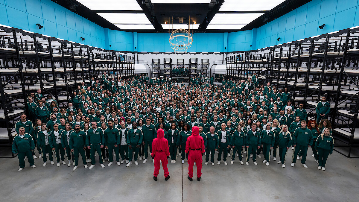 Hundreds of people in green tracksuits stand in rows inside a large, brightly lit room with bunk beds, facing two guards in pink uniforms.