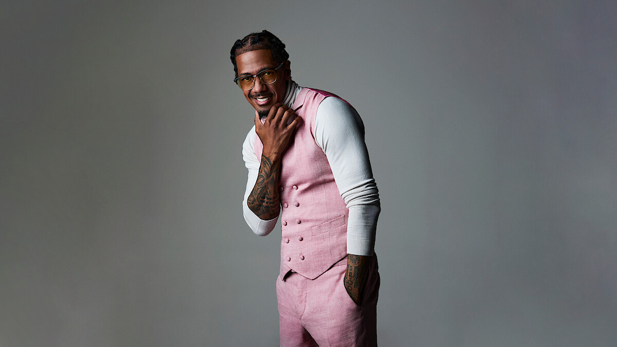 Nick Cannon in pink vest and pants over white turtleneck poses confidently in a studio with a neutral gray background, smiling while touching his chin, wearing glasses, and showing tattooed arms.