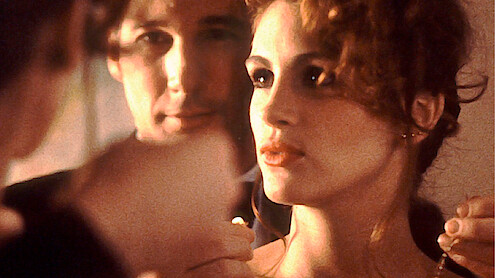 A man and woman stand closely together in a softly lit indoor setting, with the man carefully placing a necklace around the woman's neck, both reflected in a mirror, creating an intimate and elegant atmosphere.