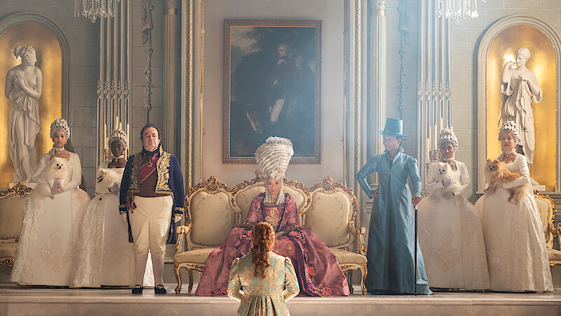 A regal court scene with a queen on an ornate throne, flanked by elaborately dressed courtiers and ladies holding small dogs, set in a lavish, gold-accented palace with classical statues and a large portrait.