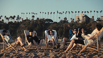j-hope, Suga, Jin, RM, Jung Kook, and Jimin relaxing on beach chairs in the sand, enjoying a sunny day. Palm trees and buildings are visible in the background.