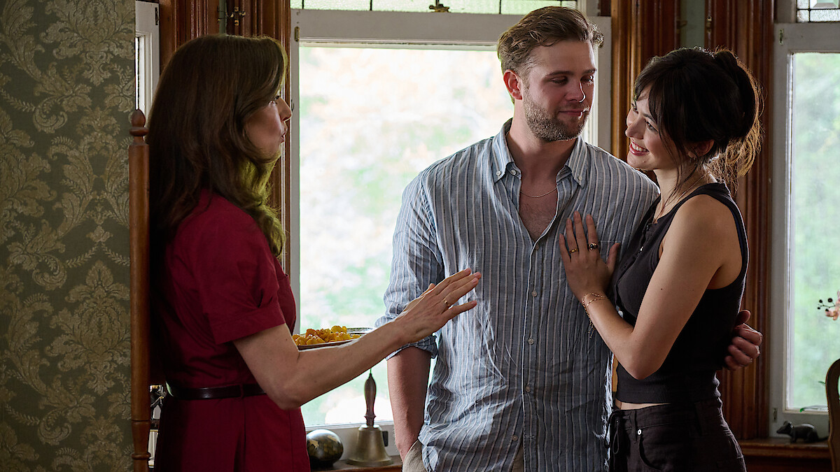 Rachel Weisz as The Protagonist, Leo Woodall as Vladimir, and Jessica Henwick as Cynthia converse in a warmly lit room with large windows. One woman in a red dress holds food, gesturing as she speaks to a smiling couple standing close together.