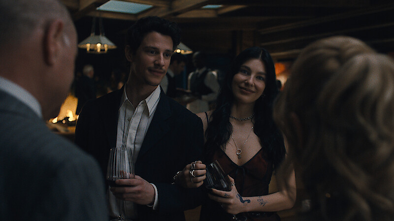 A man and woman dressed formally at a dimly lit indoor social gathering, holding drinks and smiling while conversing with other guests in an elegant setting.
