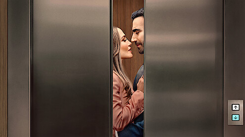 A couple standing close together inside an elevator, gazing at each other affectionately as the stainless steel elevator doors partially close, suggesting a romantic or intimate moment in a modern building setting.
