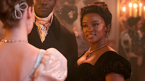 Three people dressed in elegant historical clothing at a candlelit ball, with one woman in a black gown smiling warmly.