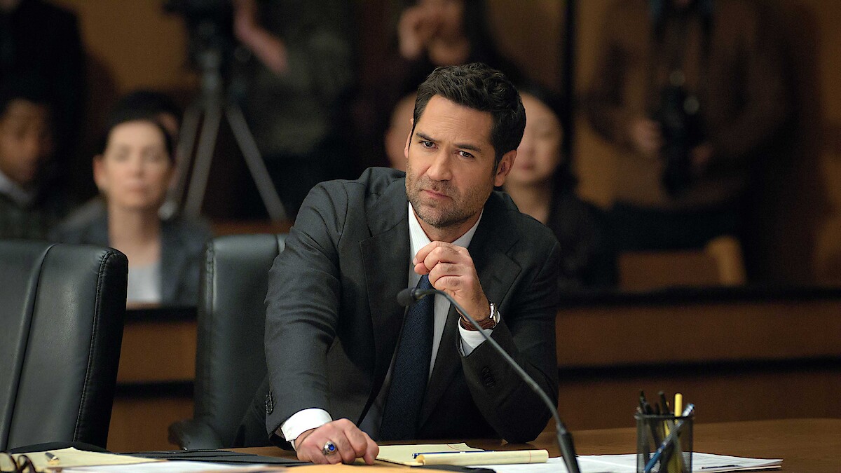 A serious-looking man in a suit sits at a courtroom table with documents, holding a microphone, while people watch in the background. The mood is tense and formal.