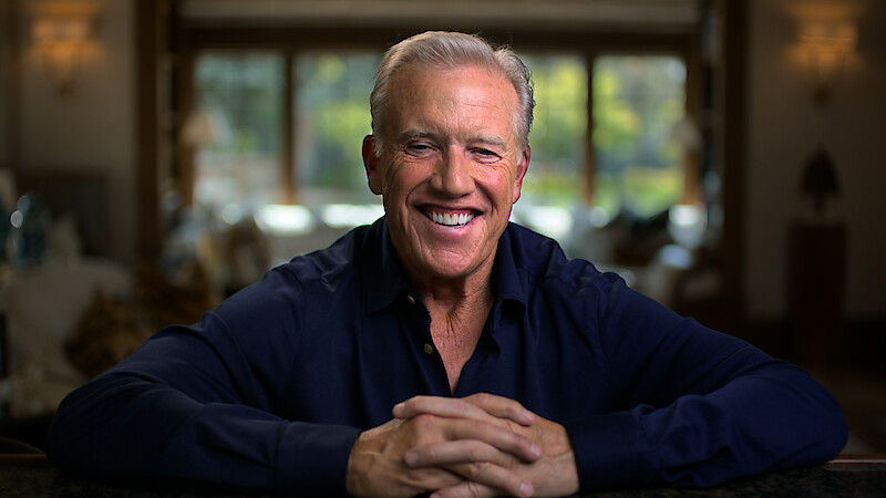 Smiling man with gray hair wearing a dark shirt sits indoors with hands folded, relaxed, in a warmly lit, blurred living room with large windows and natural light in the background.