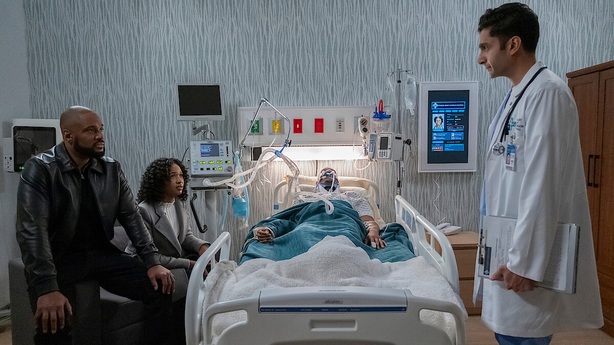 A patient lies in a hospital bed connected to medical equipment, with a man and woman seated nearby and a doctor standing at the bedside in a modern hospital room.