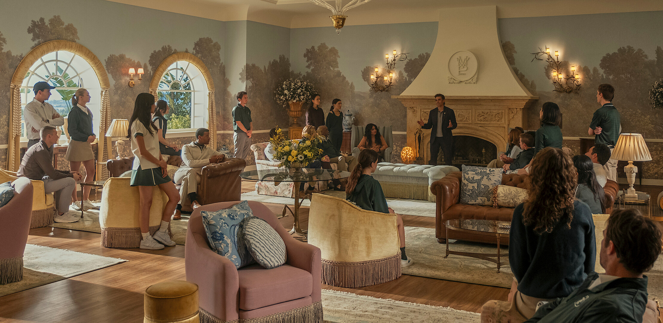 Group of employees gathered in a spacious, elegant living room with large windows, ornate fireplace, chandeliers, and stylish furniture, appearing to listen to a speaker in a formal setting.