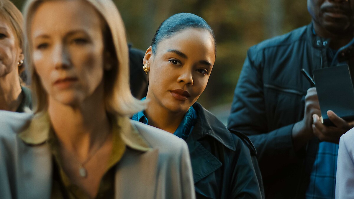 A woman in focus stands among a group of people outdoors, listening attentively, with trees in the background. The scene suggests a serious or formal gathering, possibly a press conference or official announcement.