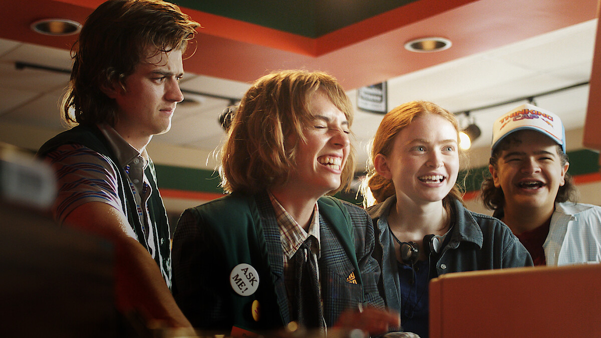 From left to right, Joe Keery, Maya Hawke, Sadie Sink, and Gaten Matarazzo stand together indoors; Keery appears serious while Hawke, Sink, and Matarazzo are smiling and laughing, visibly cheerful and engaged.