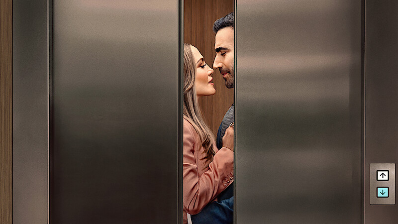 A couple standing close together inside an elevator, gazing at each other affectionately as the stainless steel elevator doors partially close, suggesting a romantic or intimate moment in a modern building setting.