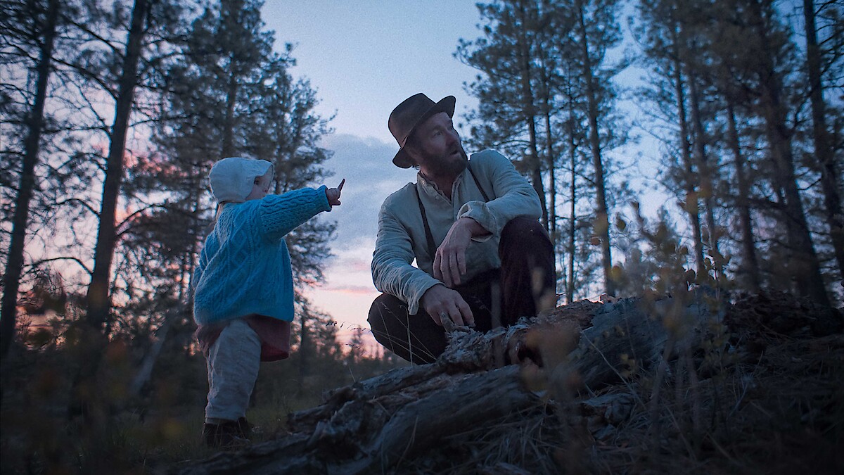 A man in a hat and a child in a blue sweater explore a forest at dusk, surrounded by tall trees. The child points at something, creating a sense of curiosity and peacefulness in the natural setting.
