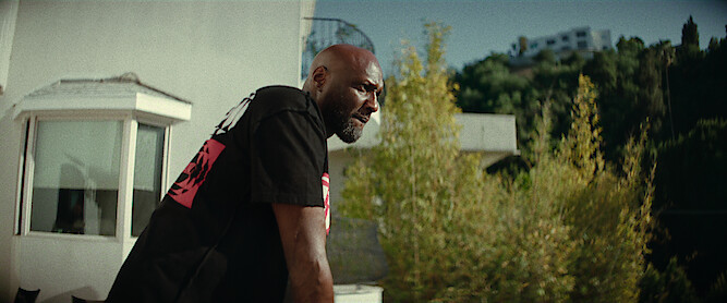 Lamar Odom wearing a black shirt stands outdoors on a balcony, leaning forward and looking into the distance; modern white buildings and greenery are visible in the background on a sunny day.