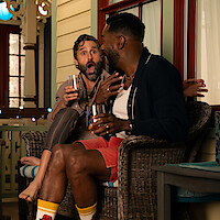Two men sitting on a porch at night, laughing and talking while holding drinks, with one looking surprised; cozy, relaxed atmosphere with decorative lights and porch furniture.