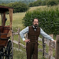 a man stands with a corn field on one side and a late 1800s carriage on the other. 